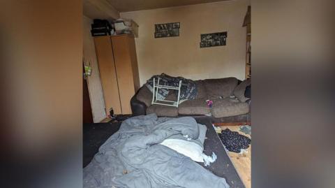 A messy house showing mess and rubbish with a dirty duvet on the floor near a brown sofa.