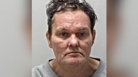 Police mugshot of Adam Saunders, who has brown unruly hair. He is photographed wearing a grey top.