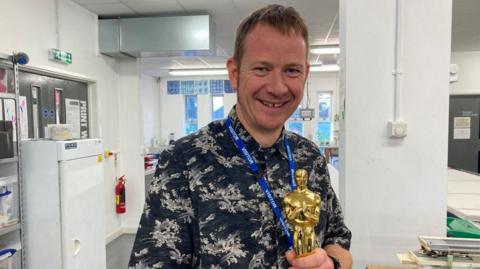 A man smiling at the camera holding an Oscar. He is wearing a dark patterned shirt. He is standing in what appears to be an office building