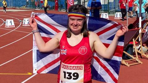A woman wearing a red sports vest holds a union jack flag in the air. She is standing on a racing track.