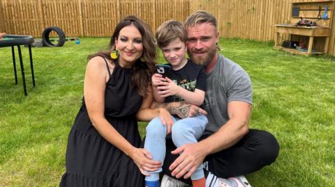 Family who use new neurodiversity centre in Dingwall - a woman with brown hair in a black dress, a man in a grey T-shirt and black trousers and a boy holding a phone with light blue jogging bottoms and a dark T-shirt. They are sitting on grass in a play area.