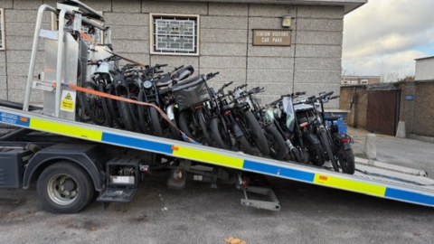 A flatbed recovery truck carrying multiple electric bikes stacked upright and secured with straps, parked beside a brick and concrete building.