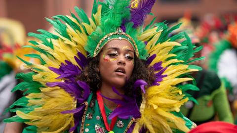 A young woman wears a large head-dress of purple, yellow and green fake feathers as she dances in a parade. She wears red, green and yellow sparkles around her eyes.