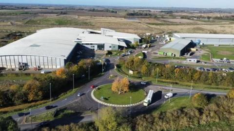 Aerial view of Waterbeach waste facility. It is situated off a busy roundabout and has a large white factory on one side of the road and a smaller light green one on the other. It is surrounded by fields.