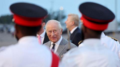 King Charles, in focus, stands between two out-of-focus soldiers wearing white uniforms and black top hats with red bases.