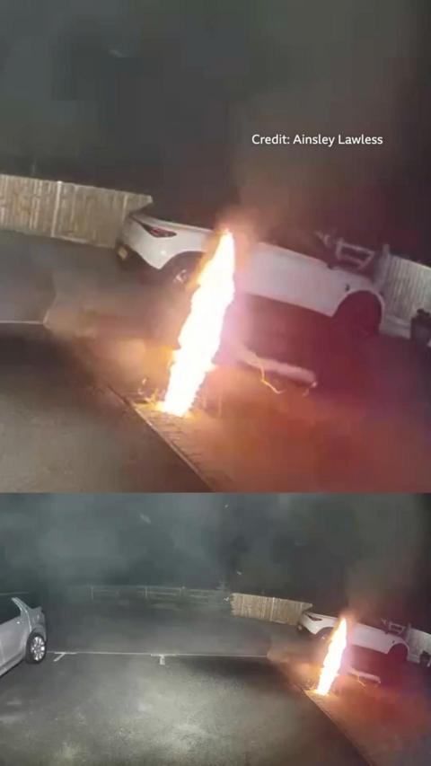 A large flame on a small patch of grass in front of a white car.