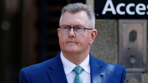 Sir Jeffrey Donaldson has short, cropped grey hair. He is wearing black-rimmed glasses and a blue suit, white shirt and a green patterned tie