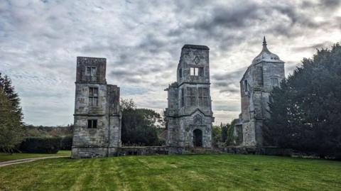 The image shows Brambletye House, three grey tower structures surrounded by trees and grass lawns. The structure is framed against a cloudy grey sky.
