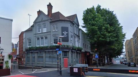 The corner of a pedestrianised street. There is a three-storey grey building with a brown roof and a chimney on the corner. There is a black rectangular sign that says "The Sly Old Fox" in white writing on the building. To the right of the building there is a row of trees and a black barrier that keeps vehicles off the street.