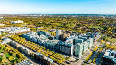 An aerial view of Milton Keynes. There are a lot of trees, office-style buildings in the foreground, and clusters of residential homes in the background.