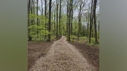 Fresh gravel path running through hardwood woodland, with a public footpath sign in the distance.