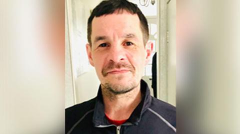 Craig Foy with brown hair and brown stubble wearing a navy zip up jumper standing in a hallway with a white front door behind him. 