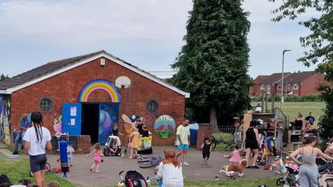 Venture Community Hub - a small red brick building with several murals painted on it, including a rainbow and two birds. Lots of children and parents can be seen in the field next to the building.