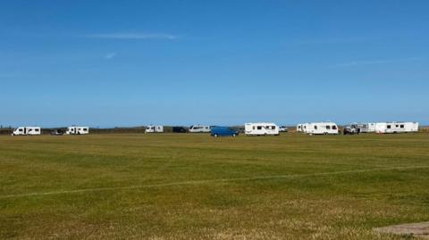 A row of white caravans have parked along the edge of a football pitch. There are tyre tracks over the pitch leading to them.