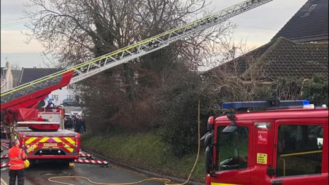 Two red fire engines on the road- with an extended metal ladder from one of them- and crews standing on the road- wearing orange jackets 