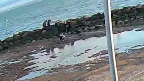 A still from CCTV footage showing a group of people near the seafront on mud or sand, with rocks immediately behind, and the sea in the background.
