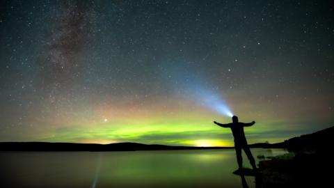 The silhouette of a man wearing a head torch in front of the green hues of the northern lights.