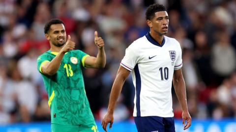 A football player wearing the green shirt of Senegal is doing a thumbs up gesture with both hands behind England's Jude Bellingham.