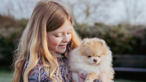 A girl with blonde hair holds a small Pomeranian puppy outdoors
