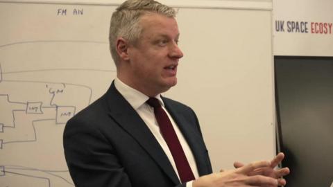 Pollard has short grey hair parted on the side. He wears a dark suit with a red tie and white shirt. He is talking to a room standing next to a whiteboard.