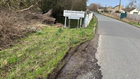 A road sign for 'High Road' and another road sign for the village of Ashton Keynes in Wiltshire positioned at the side of the village main road which has extensive road damage and crumbling tarmac at its edge. In the background are houses and the River Thames.