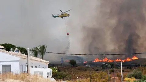 A helicopter flies over the flames and drops a bucket of water over them with a white house in the foreground and smoke rising in the background.