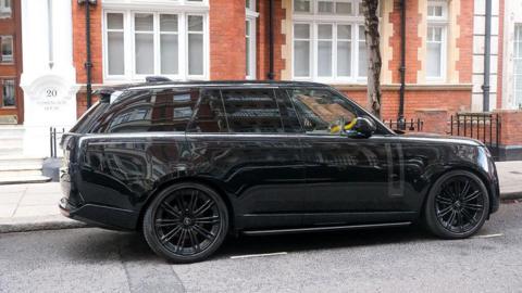 A black SUV is parked outside a brick building in London. A yellow crook lock can be seen on the wheel 
