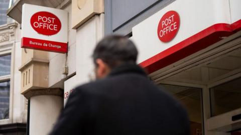 Man wearing a dark coat walks by Post Office signage in London