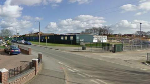 The image shows a quiet suburban street on a bright day with a partly cloudy sky. In the foreground on the left, there is a red-brick house with a grey tiled roof, bordered by a low brick wall and black metal fencing. A paved driveway or walkway runs alongside the house, and a small brick wall separates the property from the road.