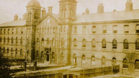 An old image showing Carlisle's Union Workhouse. The yellowed image shows the building with a main central body with a pitched roof, a tower at either side and a clock on the higher part of the facade. There are two large wings over three floors on both sides of the central body.