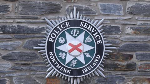 The logo of the Police Service of Northern Ireland is mounted on a stone wall at a police station. The name of the force surround the crescent. 