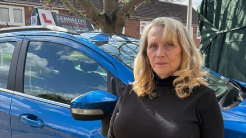 Louisa Judge stands in front of her driving instructing car which is blue. She has long blonde hair which is slightly curled. She wears a black jumper. 