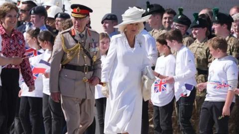 King Charles in military dress uniform and Queen Camilla in below-knee length white dress and matching hat walk past children with Union Jack flags and cadets.