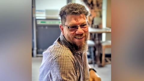 Jason Tholen is sitting and is in the middle of the image. He has short brown hair and a medium length brown beard. He is wearing a blue and red tartan shirt.