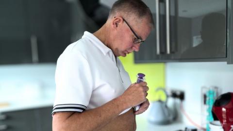 Marc Powell is wearing a white zip-up polo shirt and is in the kitchen looking at a feeding tube. 