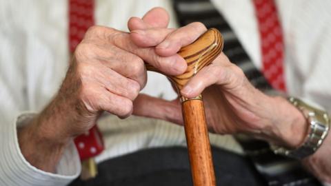 An elderly man holding a walking stick. The image is a close up of his hands. 
He is wearing a watch on this left wrist and a white shirt with red braces and a tie with black trousers.