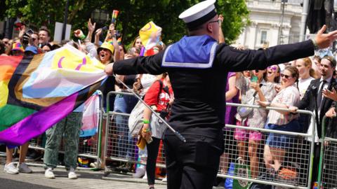 A Royal Navy sailor in uniform, carrying a LGBTQ+ flag, faces the crowd during London's Pride March.