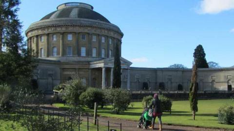 The rear of Ickworth House - a domes central rotunda with wings on either side. A woman walks with a pushchair towards the house along a gravel path with lawns either side.