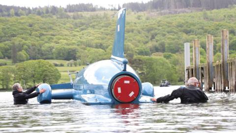 Two divers manoeuvre Bluebird next to a wooden jetty on Coniston Water. Bluebird has a tail fin and a jet engine. Trees and hills are in the background.