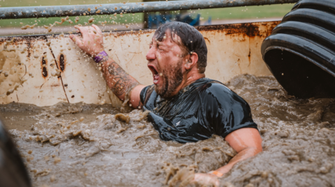A bearded man in a black t-shirt up to his neck in mud in a large container. He is grabbing onto the handrail, and his eyes are screwed shut, and his mouth is open wide with an expression of surprise