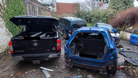 Four stripped cars in a yard. One car is blue and missing it's boot while another black car has its hood popped open