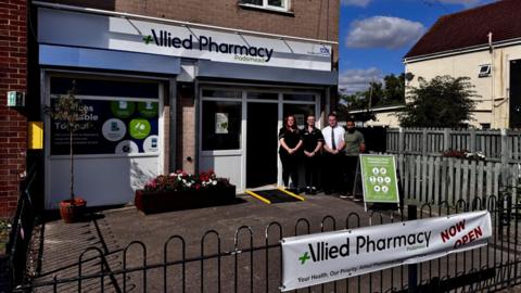 Four staff members standing outside a newly-opened pharmacy. On the railings in the foreground, a banner is hung with the words "Allied Pharmacy Now Open" visible. In the background, the four staff members stand together in front of the building which has a sign which reads "Allied Pharmacy Podsmead".