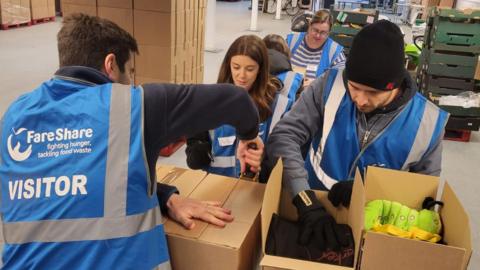 Five people are in a warehouse packing cardboard boxes. They are wearing blue high-vis vests. There are two men in the foreground and a woman who is smiling.