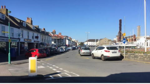 The bridge in St Sampson, card driving along a road with a row of buildings on the left and cars parked in front of them, blue skies