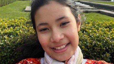 A 13-year-old girl who is pictured in a garden with a hedge behind her, smiling. She is wearing an orange outfit. 