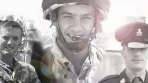 A black and white image of three soldiers. The man on the left and man in the middle are wearing army combat uniforms and the man on right is wearing a cap and ceremonial uniform with a shirt and tie. 