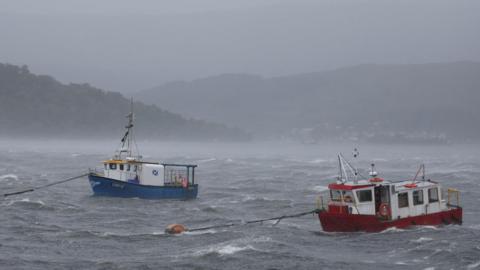 Two small creel boats, one white and blue and the other white and red, strain at their moorings at Fort William during August's Storm Floris.