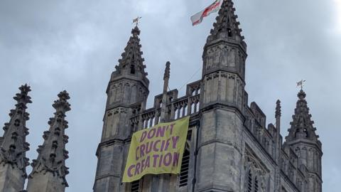 A yellow banner with the words 'don't crucify creation' written on in purple letters hangs from the pinnacle of Bath Abbey
