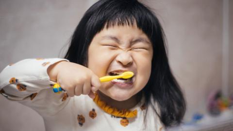 Young girl brushing teeth with yellow toothbrush, scrunching up her eyes in happiness.