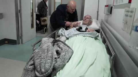 A man leans over an elderly patient lying on a trolley in a hospital corridor, with other patients and equipment visible in the background.